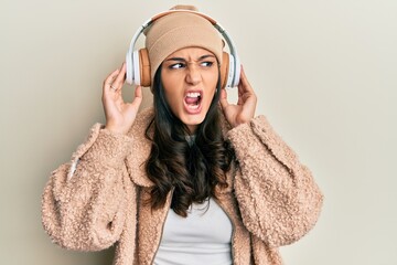 Young hispanic woman listening to music using headphones angry and mad screaming frustrated and furious, shouting with anger. rage and aggressive concept.
