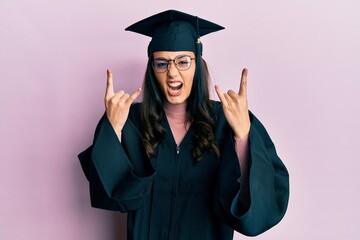 Young hispanic woman wearing graduation cap and ceremony robe shouting with crazy expression doing rock symbol with hands up. music star. heavy concept.