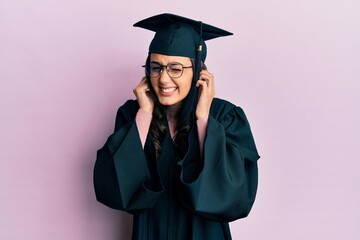 Young hispanic woman wearing graduation cap and ceremony robe covering ears with fingers with annoyed expression for the noise of loud music. deaf concept.