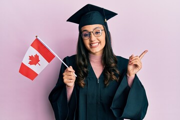 Young hispanic woman wearing graduation uniform holding canada flag smiling happy pointing with hand and finger to the side