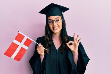 Young hispanic woman wearing graduation uniform holding denmark flag doing ok sign with fingers, smiling friendly gesturing excellent symbol