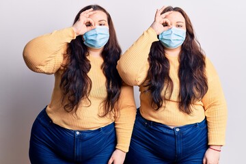 Young plus size twins wearing medical mask doing ok gesture with hand smiling, eye looking through fingers with happy face.