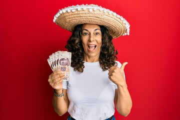 Middle age hispanic woman wearing mexican hat holding pesos banknotes pointing thumb up to the side smiling happy with open mouth
