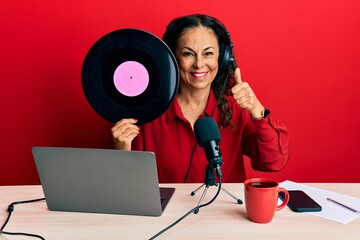 Beautiful middle age woman working at radio studio holding vinyl disc smiling happy and positive, thumb up doing excellent and approval sign