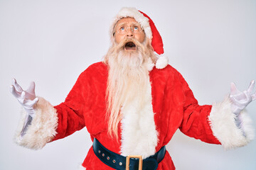 Old senior man with grey hair and long beard wearing santa claus costume with suspenders crazy and mad shouting and yelling with aggressive expression and arms raised. frustration concept.