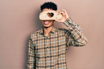 Young african american man holding bread loaf with heart shape smiling with a happy and cool smile on face. showing teeth.