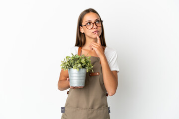 Gardener girl holding a plant over isolated white background having doubts while looking up