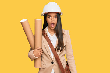 Beautiful hispanic woman holding paper blueprints scared and amazed with open mouth for surprise,...