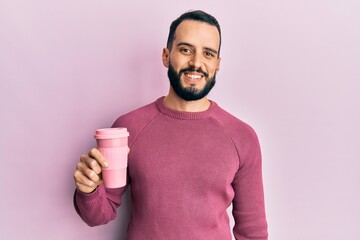 Young man with beard drinking a take away cup of coffee looking positive and happy standing and...