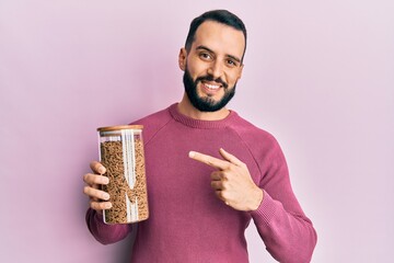 Young man with beard holding jar of healthy whole grain cereals smiling happy pointing with hand and finger