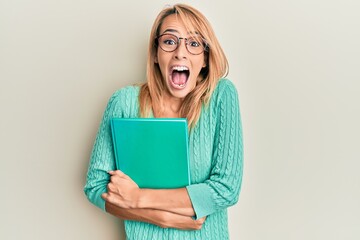 Beautiful blonde woman holding book wearing glasses celebrating crazy and amazed for success with open eyes screaming excited.