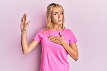 Beautiful blonde woman wearing casual pink tshirt swearing with hand on chest and open palm, making...