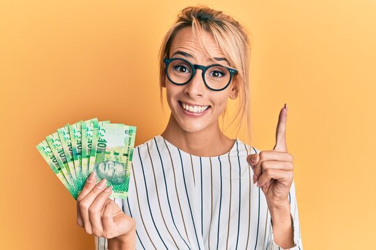 Beautiful blonde woman holding south african 10 rand banknotes smiling with an idea or question pointing finger with happy face, number one