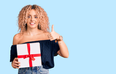 Young blonde woman with curly hair holding gift smiling happy and positive, thumb up doing excellent and approval sign