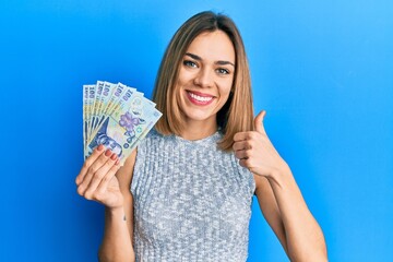 Young caucasian blonde woman holding 100 romanian leu banknotes smiling happy and positive, thumb...