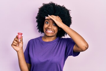 Young african american woman holding menstrual cup stressed and frustrated with hand on head,...