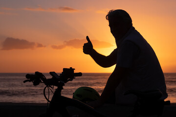 Silhouette of mature man sitting near his bicycle at the beach with a magnificent orange sunset....