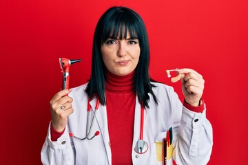 Young hispanic doctor woman holding deafness headset and otoscope skeptic and nervous, frowning...