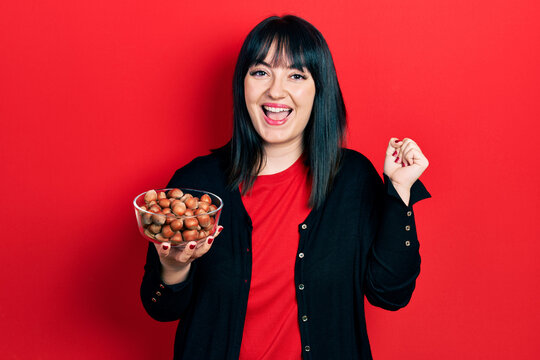 Young hispanic woman holding bowl of chestnuts screaming proud, celebrating victory and success very excited with raised arm