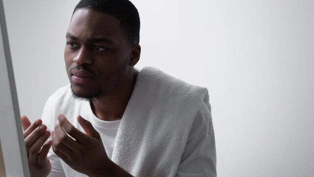 Skin problem. Acne treatment. Allergy irritation. Annoyed dissatisfied disturbed African man in white t-shirt with towel touching dry face looking in mirror.