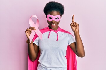 Young african american girl wearing superhero costume holding pink cancer ribbon surprised with an idea or question pointing finger with happy face, number one