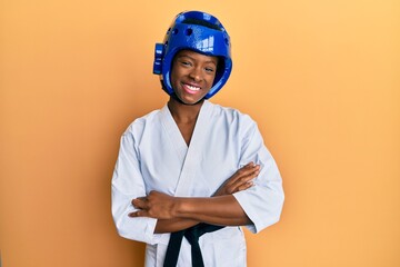 Young african american girl wearing taekwondo kimono and protection helmet happy face smiling with crossed arms looking at the camera. positive person.