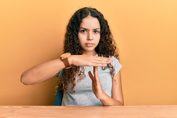 Teenager hispanic girl wearing casual clothes sitting on the table doing time out gesture with...