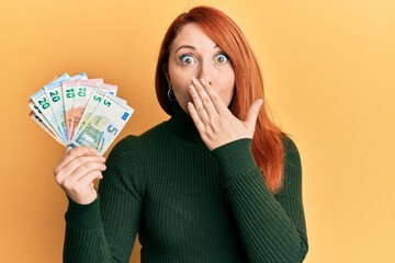 Beautiful redhead woman holding euro banknotes covering mouth with hand, shocked and afraid for mistake. surprised expression