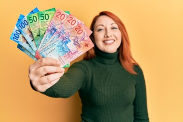 Beautiful redhead woman holding swiss franc banknotes looking positive and happy standing and smiling with a confident smile showing teeth