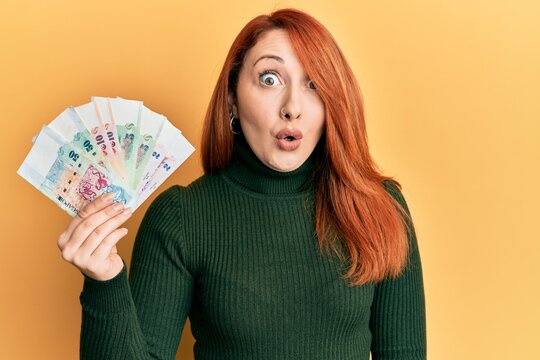 Beautiful redhead woman holding singapore dollars banknotes scared and amazed with open mouth for surprise, disbelief face
