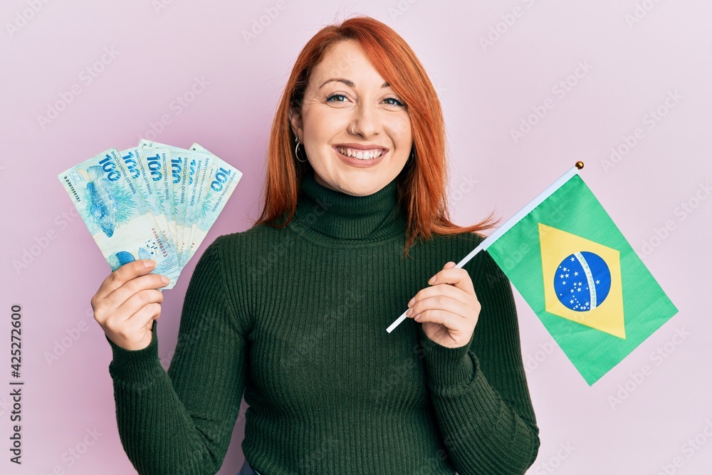 Wall mural Beautiful redhead woman holding 100 brazilian real banknotes and brazil flag smiling with a happy and cool smile on face. showing teeth.