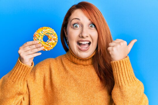 Beautiful redhead woman holding tasty colorful doughnut pointing thumb up to the side smiling happy with open mouth