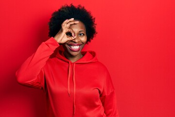 Young african american woman wearing casual sweatshirt doing ok gesture with hand smiling, eye looking through fingers with happy face.