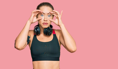 Beautiful blonde woman wearing gym clothes and using headphones trying to open eyes with fingers, sleepy and tired for morning fatigue