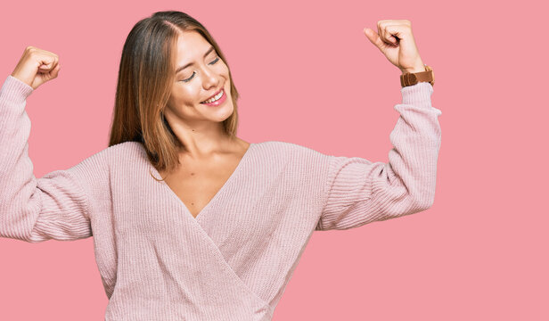 Beautiful blonde woman wearing casual winter pink sweater showing arms muscles smiling proud. fitness concept.