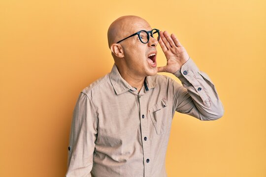 Middle age bald man wearing casual clothes and glasses shouting and screaming loud to side with hand on mouth. communication concept.