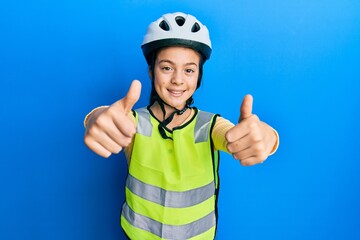 Beautiful brunette little girl wearing bike helmet and reflective vest approving doing positive gesture with hand, thumbs up smiling and happy for success. winner gesture.