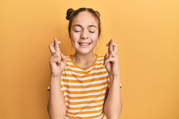 Beautiful brunette little girl wearing casual striped t shirt gesturing finger crossed smiling with hope and eyes closed. luck and superstitious concept.