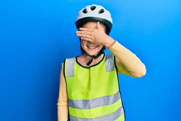 Beautiful brunette little girl wearing bike helmet and reflective vest smiling and laughing with hand on face covering eyes for surprise. blind concept.