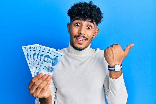 Young arab handsome man holding 10 yuan chinese banknotes pointing thumb up to the side smiling happy with open mouth