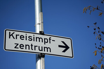 Fototapeta premium Sign on Column saying: District Vaccination Center (germany: Kreisimpfzentrum). branches of tree Isolated on blue sky. Germany, Esslingen, Zell.
