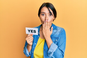 Beautiful young woman with short hair holding yes reminder covering mouth with hand, shocked and...