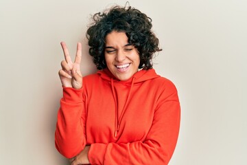 Young hispanic woman with curly hair wearing casual sweatshirt smiling with happy face winking at...