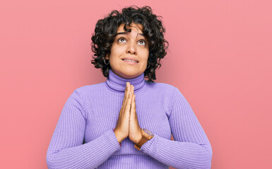 Young hispanic woman with curly hair wearing casual clothes begging and praying with hands together...