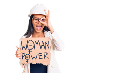 Young beautiful latin girl wearing architect hardhat holding woman power banner smiling happy doing ok sign with hand on eye looking through fingers