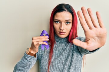 Young caucasian woman holding purple ribbon awareness with open hand doing stop sign with serious...