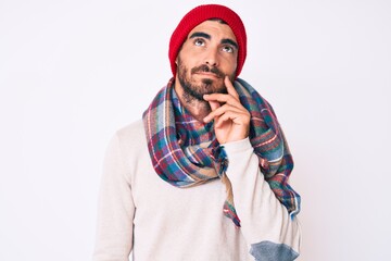 Handsome young man with curly hair and bear wearing a winter sweater, scarf and wool hat thinking...