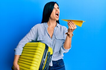 Young caucasian woman holding cabin bag and paper airplane angry and mad screaming frustrated and...