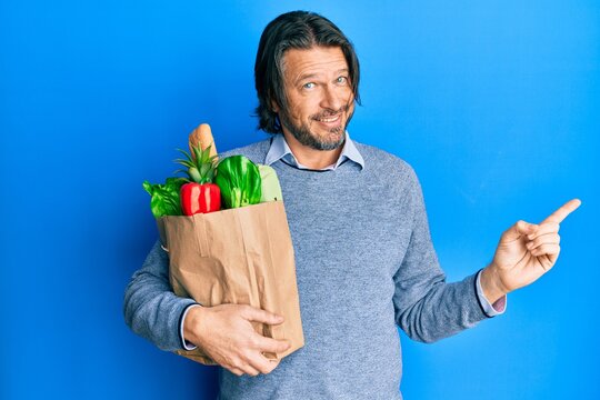 Middle age handsome man holding paper bag with groceries smiling happy pointing with hand and finger to the side