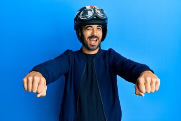Young hispanic man holding motorcycle helmet winking looking at the camera with sexy expression,...